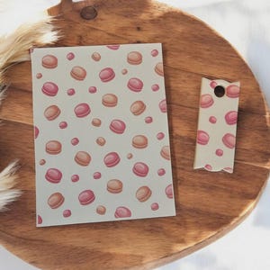 May include: A stationery set featuring a repeating pattern of pink and peach macarons on a light beige background. The set includes a rectangular card and a tag with a hole, both with the same design. The items are displayed on a wooden surface.