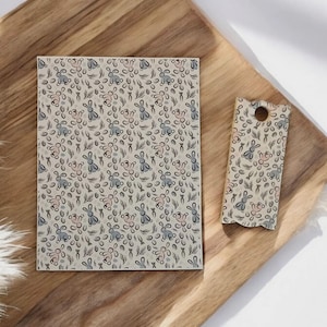 May include: A rectangular card and a tag with a repeating pattern of bunnies, carrots, and leaves in blue, pink, and black on a beige background. The items are on a wooden tray.