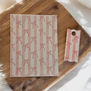 Puede incluir: Una tarjeta rectangular y una etiqueta con un patrón de lazo y rayas rosas. El fondo es de color beige claro. La tarjeta y la etiqueta están sobre una bandeja de madera.