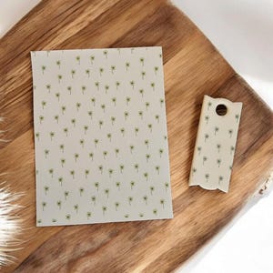 May include: A rectangular notepad and a small, decorative cutting board with a hole, both featuring a repeating pattern of small yellow flowers on a cream background. The items rest on a wooden surface.