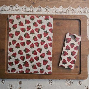 May include: A wooden tray holds a notebook and a phone case, both adorned with a repeating pattern of red strawberries on a white background. The tray is a warm brown color, and the scene is set on a white lace tablecloth.