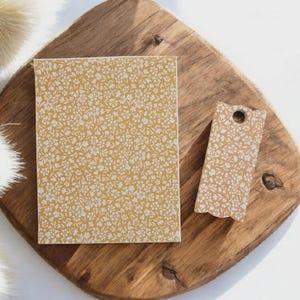 May include: A rectangular notepad and a tag with a floral pattern in white on a mustard yellow background. The notepad and tag are on a wooden cutting board. The tag has a hole at the top.
