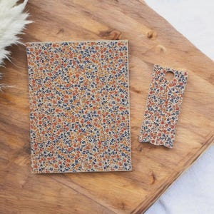 Puede incluir: Un cuaderno y un marcapáginas con estampado floral descansan sobre una bandeja de madera. El cuaderno es rectangular y el marcapáginas tiene un agujero en la parte superior. El diseño floral presenta pequeñas flores naranjas, azules y blancas.