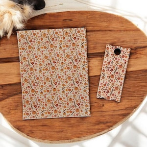 May include: A notebook and a bookmark with a fall-themed pattern of pumpkins, flowers, and leaves in orange, red, and brown on a cream background. The items are on a wooden tray.