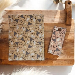 May include: A notebook and bookmark featuring a repeating pattern of owls, eyeglasses, lightning bolts, wizard hats, and brooms in shades of gray and white on a tan background. The bookmark is a lighter shade of tan.