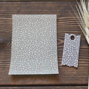 Puede incluir: Una hoja rectangular y una etiqueta con borde festoneado, ambas en gris claro con un patrón de lunares blancos. Un manojo de espigas de trigo sobre una superficie de madera oscura.