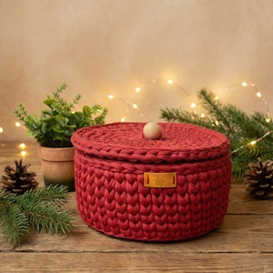 May include: A round, red crocheted storage basket with a matching lid and a wooden knob. A small, rectangular leather tag with a snowflake design is attached to the front. The basket is on a wooden surface, with greenery and lights in the background.