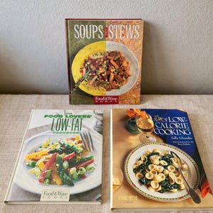 Puede incluir: Tres libros de cocina con cubiertas coloridas. El libro superior se titula "Soups & Stews" con una imagen dividida de sopa. El libro del medio es "The Food Lovers' Low-Fat Cookbook". El libro inferior es "The Art of Low-Calorie Cooking".
