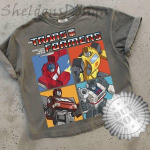 May include: Grey t-shirt with a vibrant Transformers graphic. The design features the Transformers logo and the slogan "More Than Meets The Eye." The shirt displays four different Transformers characters in action poses.