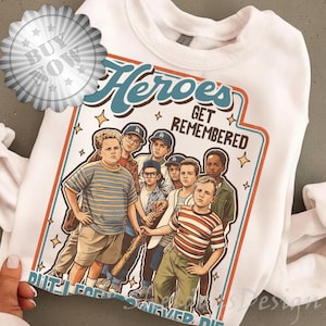 May include: White sweatshirt featuring a graphic of a group of boys with the text "Heroes Get Remembered, Legends Never Die." The illustration has a vintage style with a baseball bat and stars.