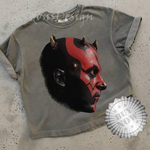 May include: Gray t-shirt featuring a graphic of a character's face with red and black details and horns. The shirt has rolled-up sleeves and a "Buy Now" sticker.