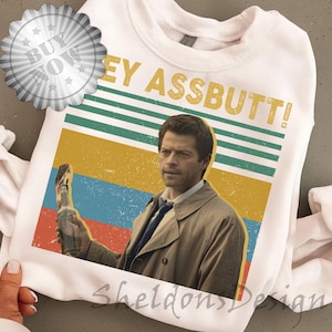 Könnte beinhalten: Weißes Sweatshirt mit dem Text "HEY ASSBUTT!" über einer Grafik eines Mannes, der eine Flasche hält. Die Grafik hat ein Retro-Farbschema mit roten, blauen, gelben und grünen Streifen. Auf der linken oberen Ecke befindet sich ein "Jetzt kaufen"-Aufkleber.