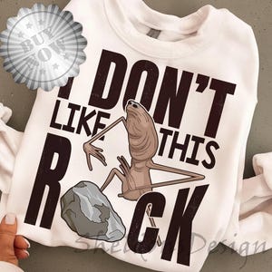 Puede incluir: Sudadera blanca con la frase "I DON'T LIKE THIS ROCK" en letras marrón oscuro. El diseño presenta una criatura alienígena de dibujos animados y una roca gris. Una pegatina de "Buy Now" está en la esquina superior izquierda.