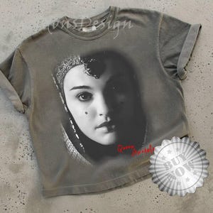 May include: Gray t-shirt featuring a black and white portrait of a woman with a headpiece. The text "Queen Amidala" is printed in red. The shirt has rolled-up sleeves and a "Buy Now" sticker.