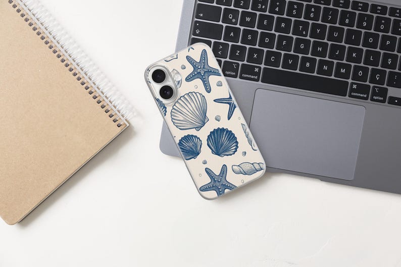 May include: A phone case with a cream background featuring a blue seashell and starfish pattern. The phone case is resting on a laptop, with a spiral notebook to the left. The laptop keyboard is visible.
