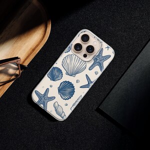 May include: A white phone case with a blue seashell and starfish pattern. The case is lying on a black surface next to a wooden tray, glasses, and a black notebook.