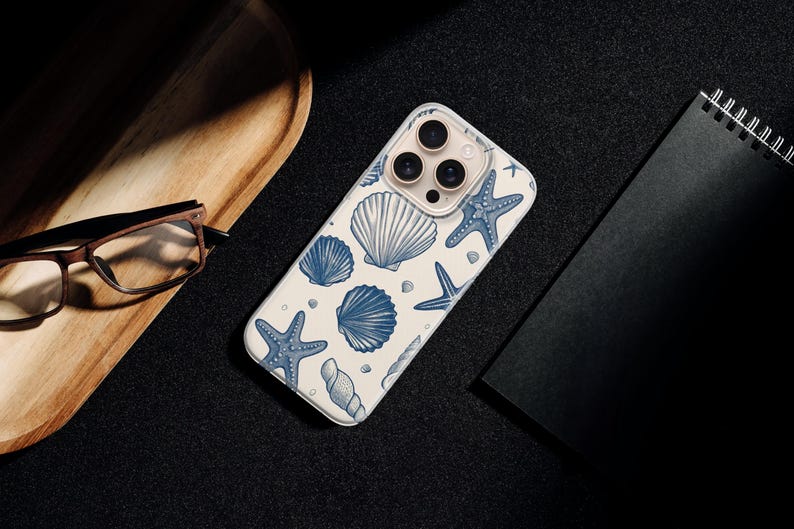May include: A white phone case with a blue seashell and starfish pattern. The case is lying on a black surface next to a wooden tray, glasses, and a black notebook.