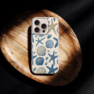 May include: A phone case featuring a repeating pattern of blue starfish, seashells, and other marine life on a cream background. The case is shown on a wooden tray against a dark background.