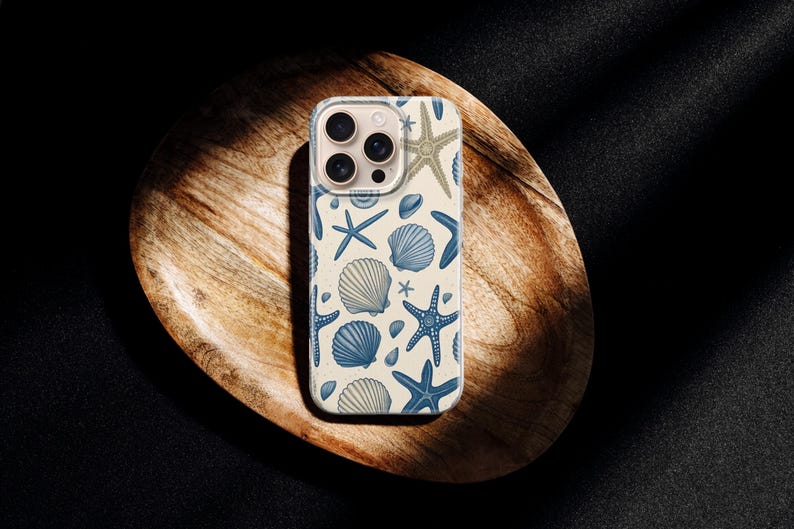 May include: A phone case featuring a repeating pattern of blue starfish, seashells, and other marine life on a cream background. The case is shown on a wooden tray against a dark background.