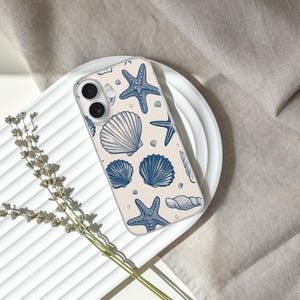 May include: A clear phone case featuring a pattern of blue seashells and starfish on a cream background. The case is resting on a white plate, with a sprig of dried flowers and a neutral-colored fabric in the background. The design is nautical.