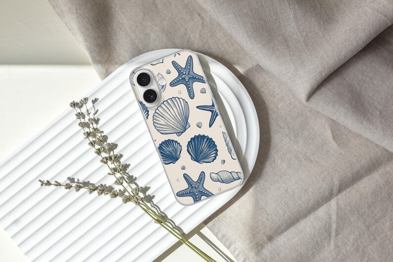 May include: A clear phone case featuring a pattern of blue seashells and starfish on a cream background. The case is resting on a white plate, with a sprig of dried flowers and a neutral-colored fabric in the background. The design is nautical.