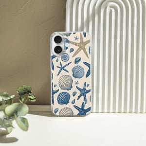 May include: A clear phone case featuring a pattern of blue starfish, seashells, and sand-colored starfish on a cream background. The case is displayed next to a white, arched decorative piece and a sprig of green leaves.