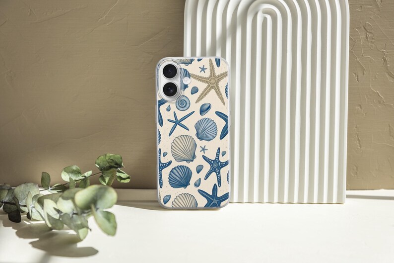 May include: A clear phone case featuring a pattern of blue starfish, seashells, and sand-colored starfish on a cream background. The case is displayed next to a white, arched decorative piece and a sprig of green leaves.