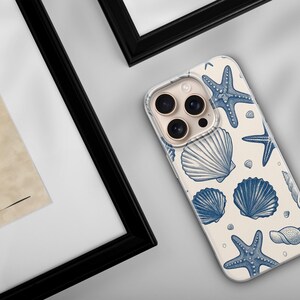 May include: A phone case with a cream background and a pattern of blue seashells and starfish. The phone case is angled, showing the camera lenses. The image also includes framed artwork.