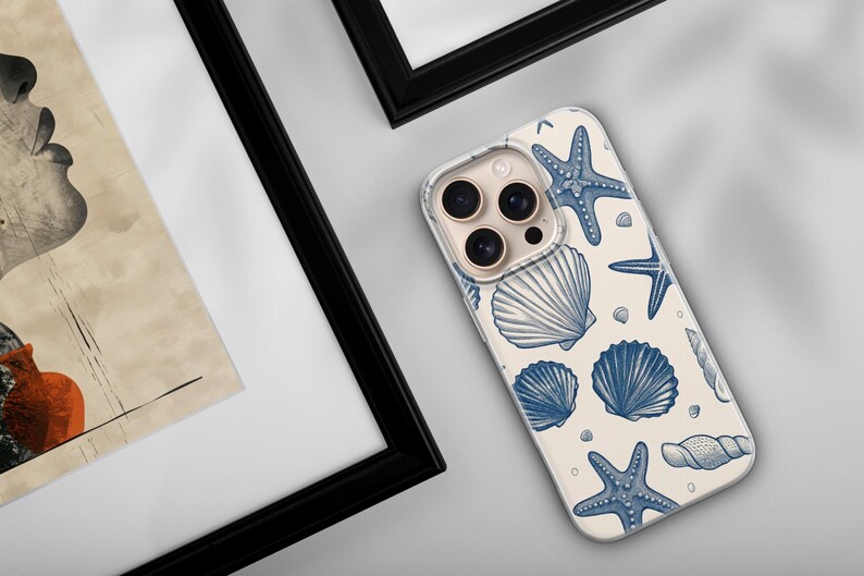 May include: A phone case with a cream background and a pattern of blue seashells and starfish. The phone case is angled, showing the camera lenses. The image also includes framed artwork.