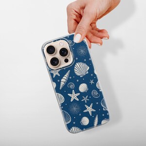 May include: A navy blue phone case featuring a pattern of white seashells, starfish, and compass designs. The case is held by a hand against a white background. The design evokes a nautical or beach theme.