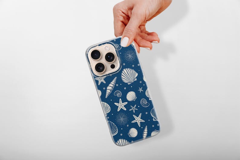 May include: A navy blue phone case featuring a pattern of white seashells, starfish, and compass designs. The case is held by a hand against a white background. The design evokes a nautical or beach theme.