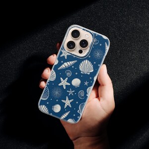 May include: A navy blue phone case featuring a repeating pattern of white seashells, starfish, and nautical elements. The case is held in a hand against a dark background. The design includes various shell types and starfishes.