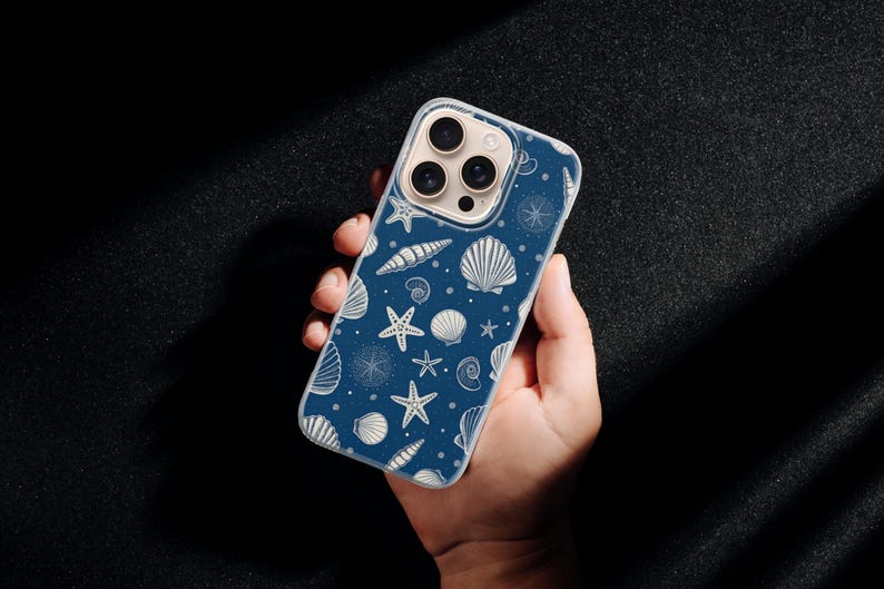 May include: A navy blue phone case featuring a repeating pattern of white seashells, starfish, and nautical elements. The case is held in a hand against a dark background. The design includes various shell types and starfishes.