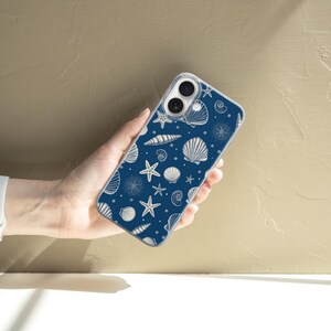May include: A navy blue phone case featuring a white seashell pattern. The design includes various seashells and starfish. The phone case is held in a hand, showcasing the detailed nautical design.