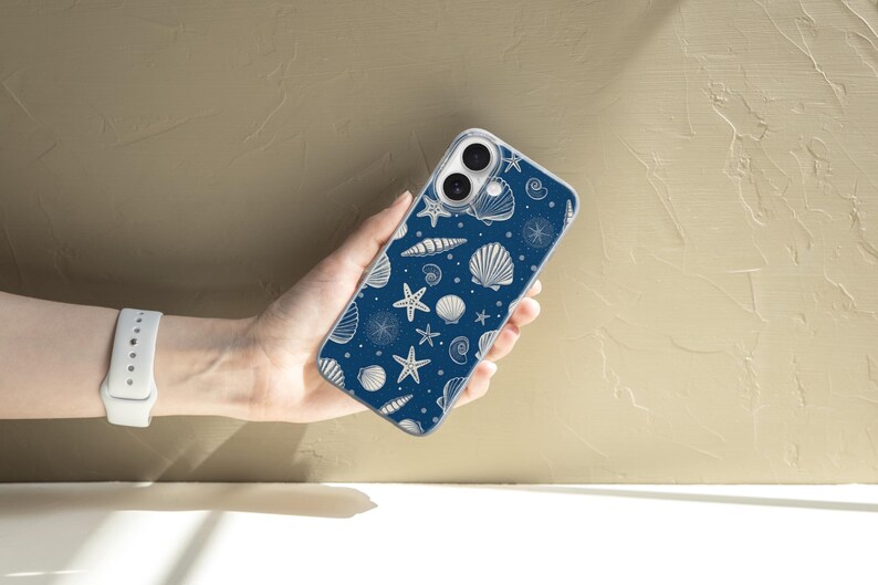 May include: A navy blue phone case featuring a white seashell pattern. The design includes various seashells and starfish. The phone case is held in a hand, showcasing the detailed nautical design.
