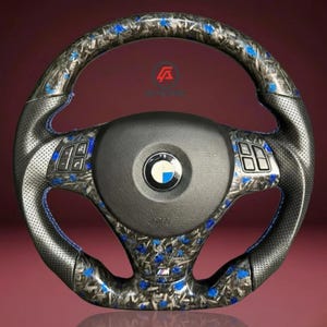 May include: A custom BMW steering wheel with a black and blue floral pattern. The wheel has perforated leather grips, blue stitching, and integrated controls. The BMW logo is centered on the airbag cover.