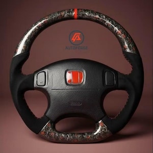 May include: Black steering wheel with a red accent stripe at the top. The wheel has a carbon fiber pattern on the grip and a red logo. The center of the wheel has a red emblem. The text "AUTOFORGE" is visible.