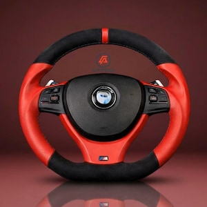 May include: A red and black steering wheel with a perforated leather grip. The wheel has a black center with a silver emblem and integrated controls. The design includes a red stripe at the top and the letters "M" and "W" at the bottom.