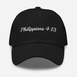 May include: A black baseball cap featuring the white embroidered text Philippians 4:13 in a cursive script font across the front panel. The hat has a structured crown, a curved brim, and a button at the top centre of the cap's dome.