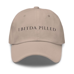 May include: A beige baseball cap featuring the black text "EBITDA PILLED" across the front. The cap has a curved peak and a button at the crown. A casual hat.