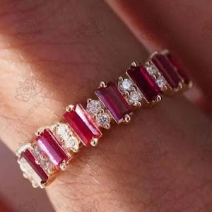 May include: A gold band ring featuring an alternating pattern of rectangular baguette-cut red rubies and pairs of small round white diamonds. The stones are set in a continuous row along the top of the metal band, creating a vibrant, multi-stone design.
