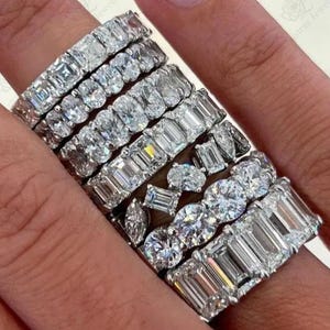 May include: A collection of eight silver-toned metal eternity bands featuring various clear gemstone cuts including emerald, oval, round, and marquise shapes. Each ring is set with multiple stones arranged in a continuous row around the band.