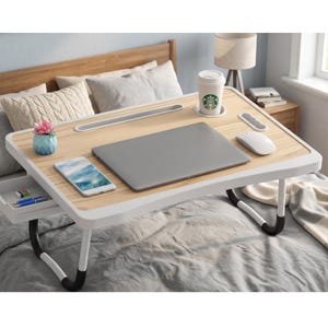May include: A light wood-tone laptop desk with white trim and folding legs. A gray laptop, white mouse, and smartphone rest on the desk. A small drawer holds pens, and a cup of coffee and a small plant are also on the desk.