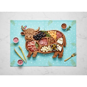 May include: A wooden serving tray shaped like a Highland cow, filled with various snacks. The tray holds grapes, cheese, salami, olives, nuts, and prosciutto. A small bird sits on the cow's head. The tray is on a blue dotted placemat with a fork and tongs.