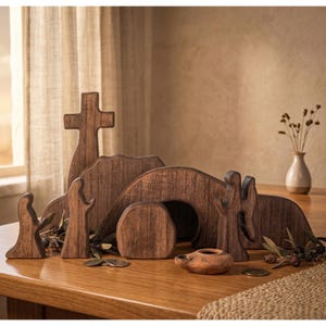 May include: Wooden Easter scene featuring a cross, tomb, and figures. The set includes a cross, a tomb with an arched opening, and two figures. A small, terracotta oil lamp and sprigs of greenery are also included. The scene is set on a wooden table.