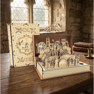 May include: A wooden laser-cut book with a religious theme. The open book reveals a detailed diorama of a biblical scene, with figures, buildings, and trees. The closed book cover features the word "HOLY" and other text.