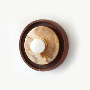 Puede incluir: Aplique de pared redondo y moderno, con un anillo exterior de madera oscura, un disco central de piedra beige y marrón, y una pequeña bombilla esférica blanca. La lámpara tiene una base de metal dorado y está montada en una pared blanca.