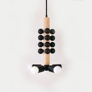 Puede incluir: Lámpara colgante moderna con una columna central de madera natural, esferas negras y brazos negros que sostienen bombillas blancas. La lámpara cuelga de un cable negro sobre un fondo blanco. Diseño geométrico.