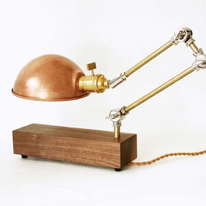 May include: A copper-colored desk lamp with a domed shade and adjustable arm. The lamp features a wooden base and brass accents. The cord is brown. The lamp is designed in an industrial style.