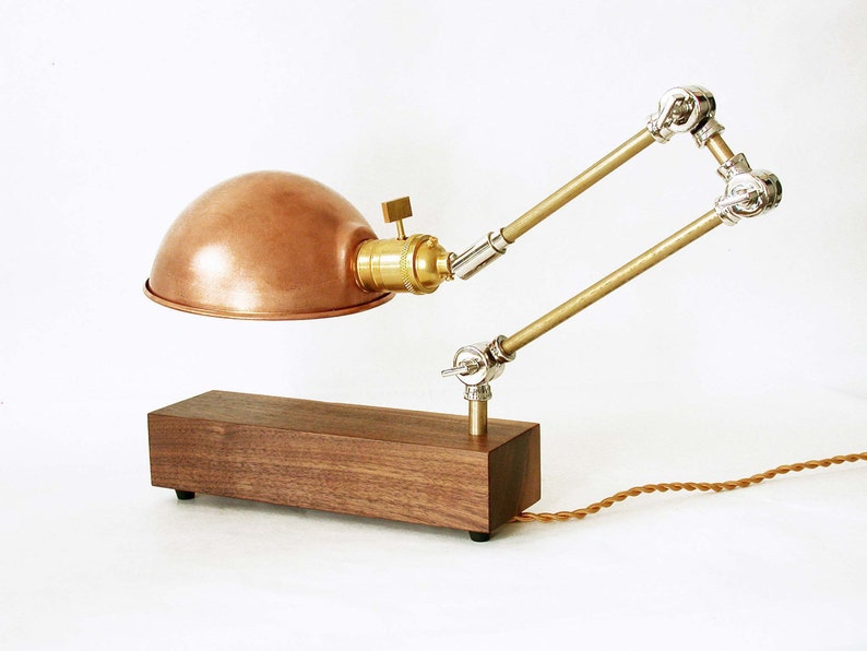 May include: A copper-colored desk lamp with a domed shade and adjustable arm. The lamp features a wooden base and brass accents. The cord is brown. The lamp is designed in an industrial style.